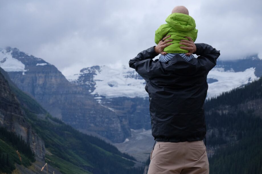 mężczyzna w czarnej kurtce trzymający na barkach dziecko w zielonej kurtce w górach